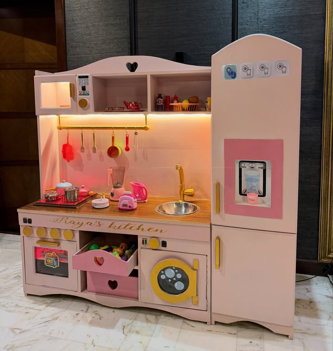 Pink wooden toy play kitchen with miniature appliances and cooking accessories for children.