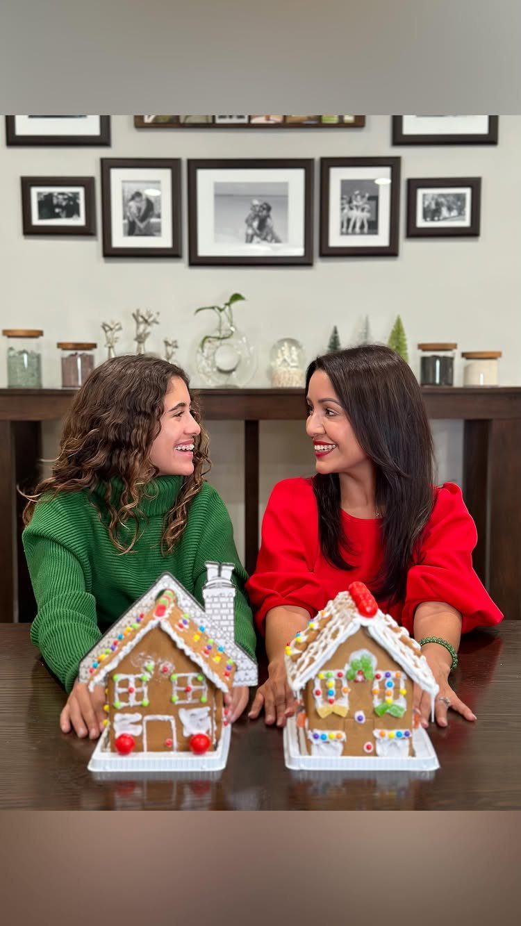Two women smiling behind two decorated gingerbread houses on a wooden table.