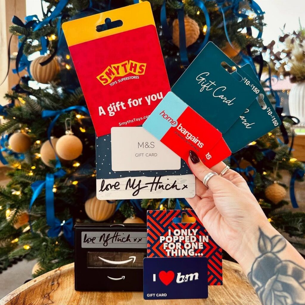 Hand holding various colorful retail gift cards in front of a decorated Christmas tree.