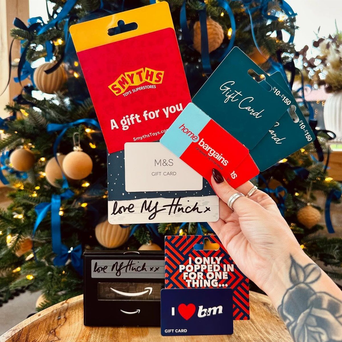 Hand holding various colorful retail gift cards in front of a decorated Christmas tree.