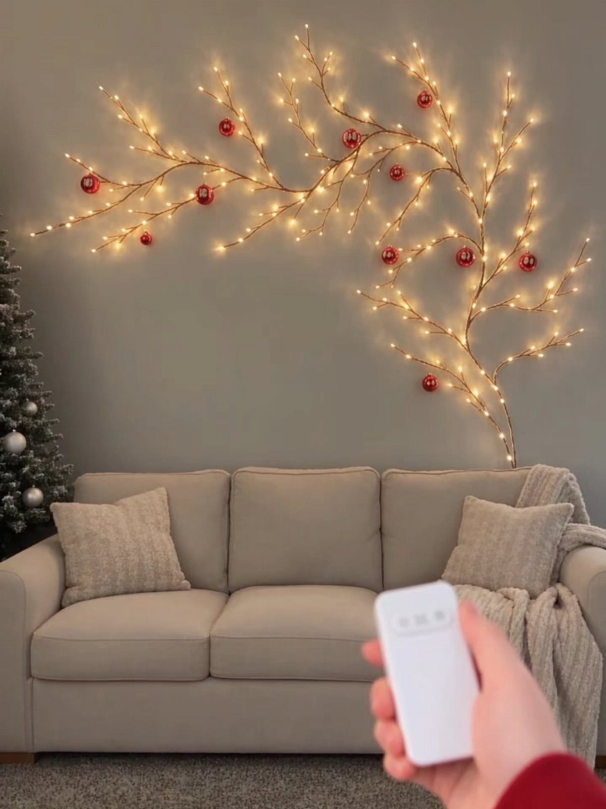 Remote control pointing to a branch-shaped wall light with red ornaments and warm glow.