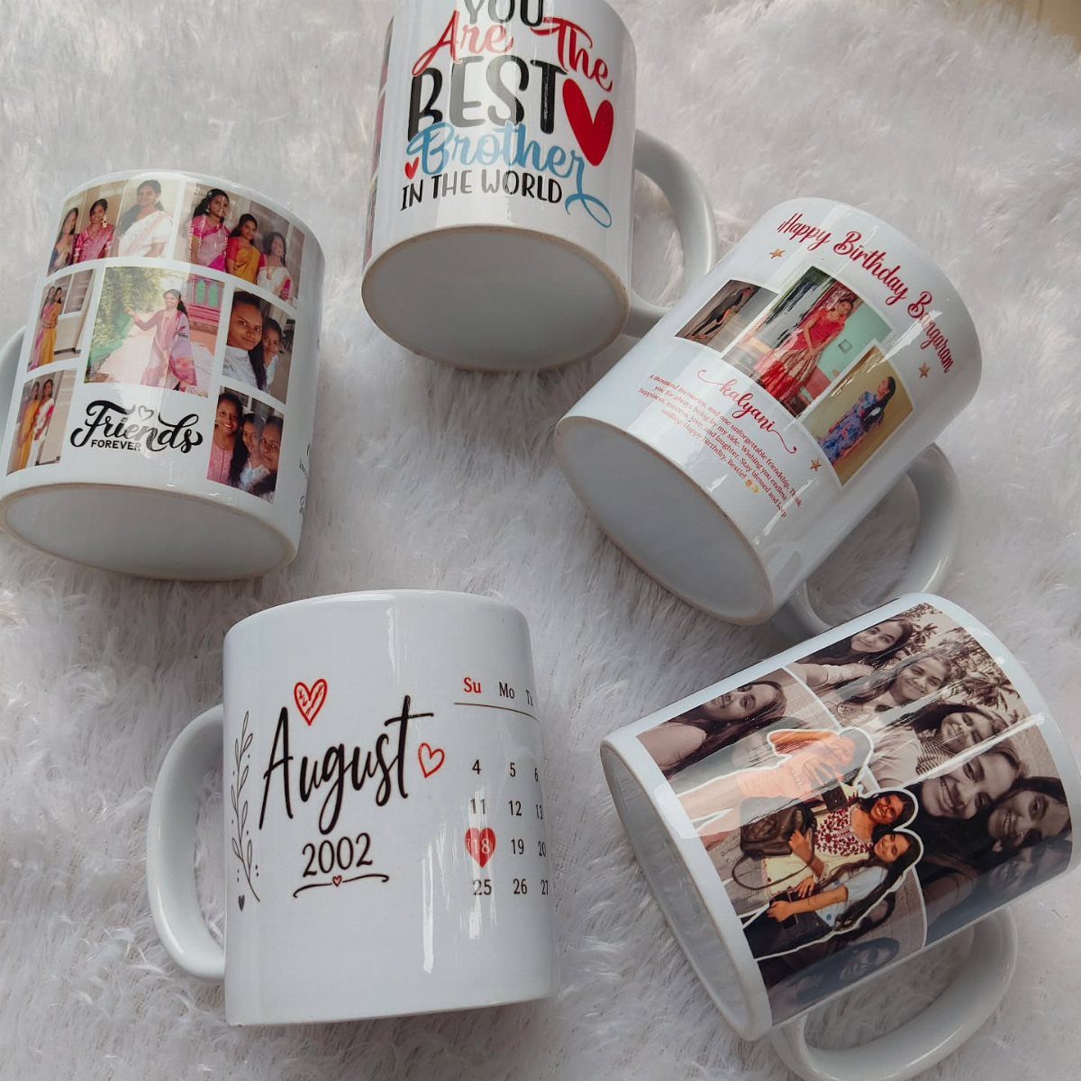 Five white ceramic mugs with personalized photos, dates, and messages on fluffy white fabric.