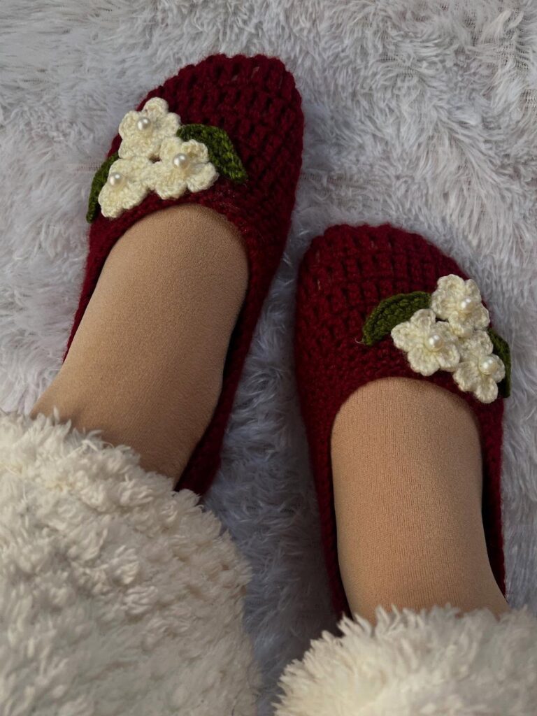 Burgundy crochet slipper socks with white flower accents on a white fluffy rug