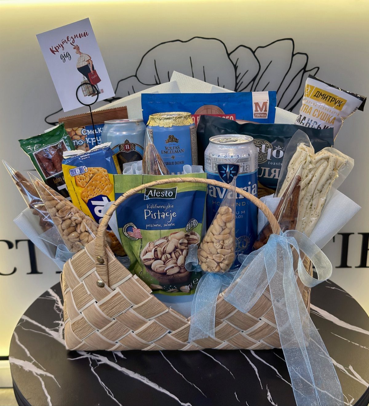 Woven gift basket filled with beer, pistachios, and snacks on a marble table.
