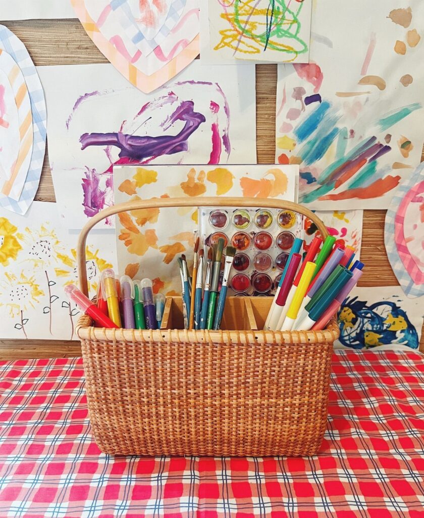 Wicker basket filled with markers, paintbrushes, and watercolors on a checkered tablecloth.