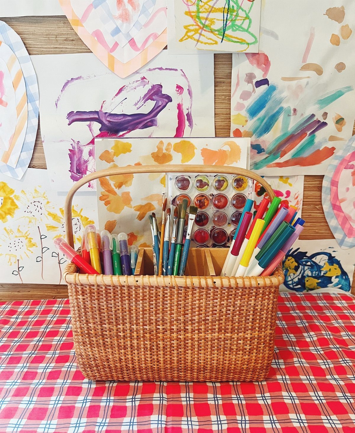 Wicker basket filled with markers, paintbrushes, and watercolors on a checkered tablecloth.