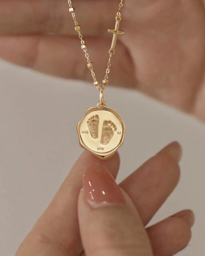Gold necklace with a cross and a pendant engraved with baby footprints.