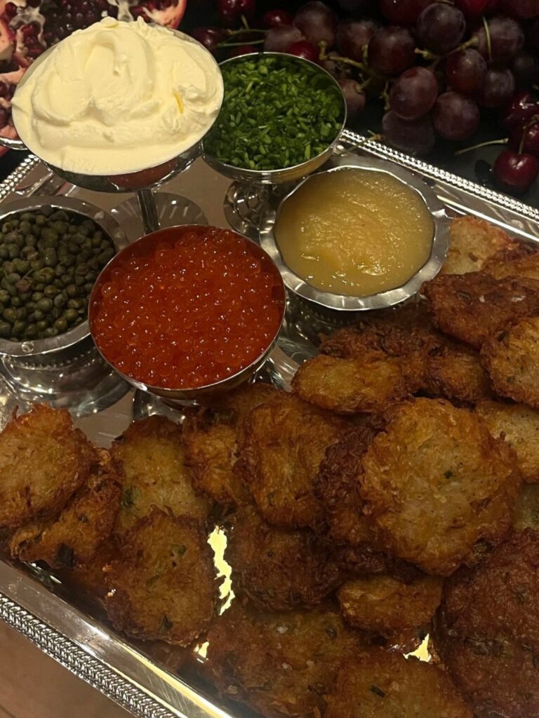 Fried potato latkes on a silver platter with various dipping sauces and grapes.