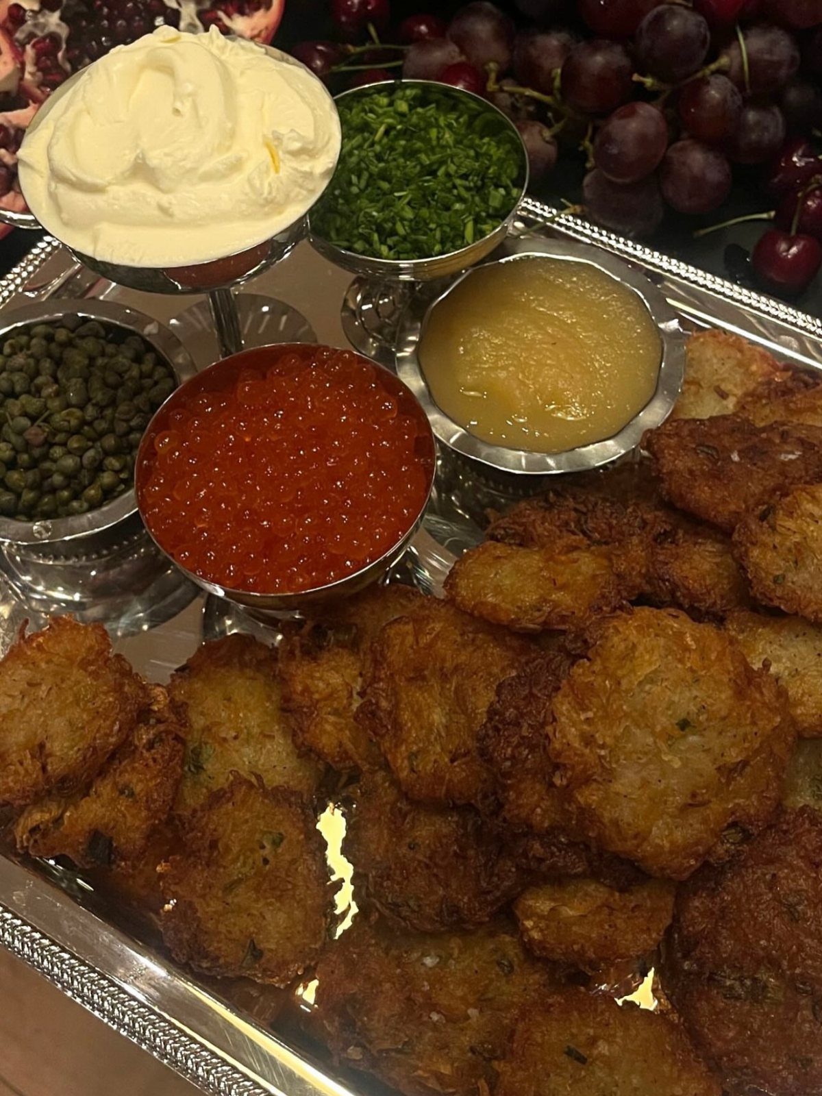 Fried potato latkes on a silver platter with various dipping sauces and grapes.
